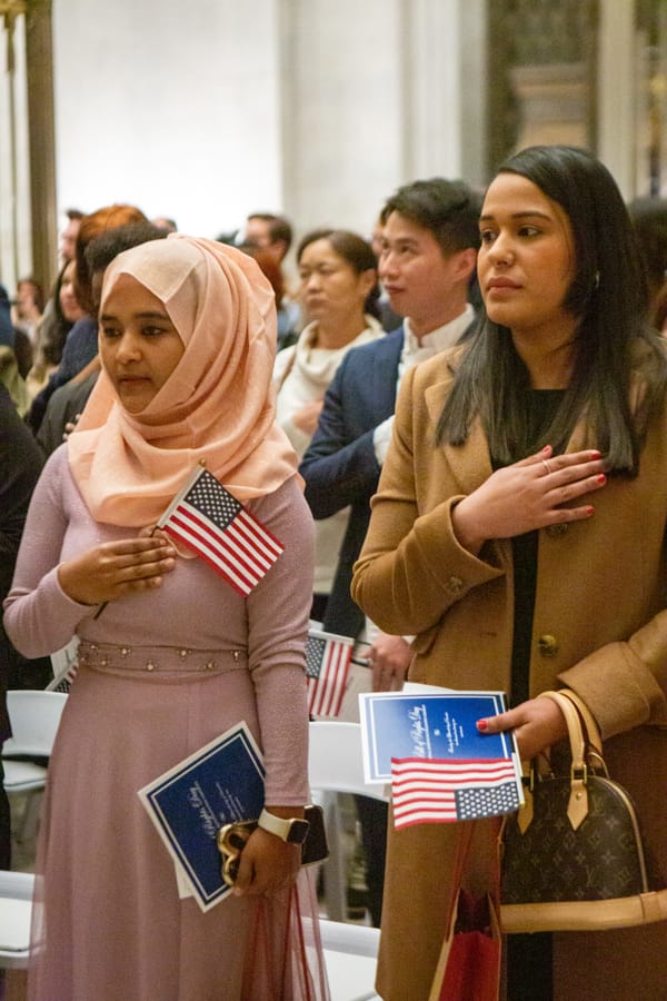 Two women holding American flags and naturalization certificates put their hands over their hearts.  